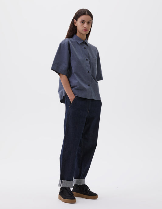 Full body image, front view of model wearing greyish blue short sleeve shirt, with elbow length sleeves, patch chest pocket and vent split hem, finished with black corozo brace buttons. Styled with indigo blue jeans and black nubuck leather trainers.