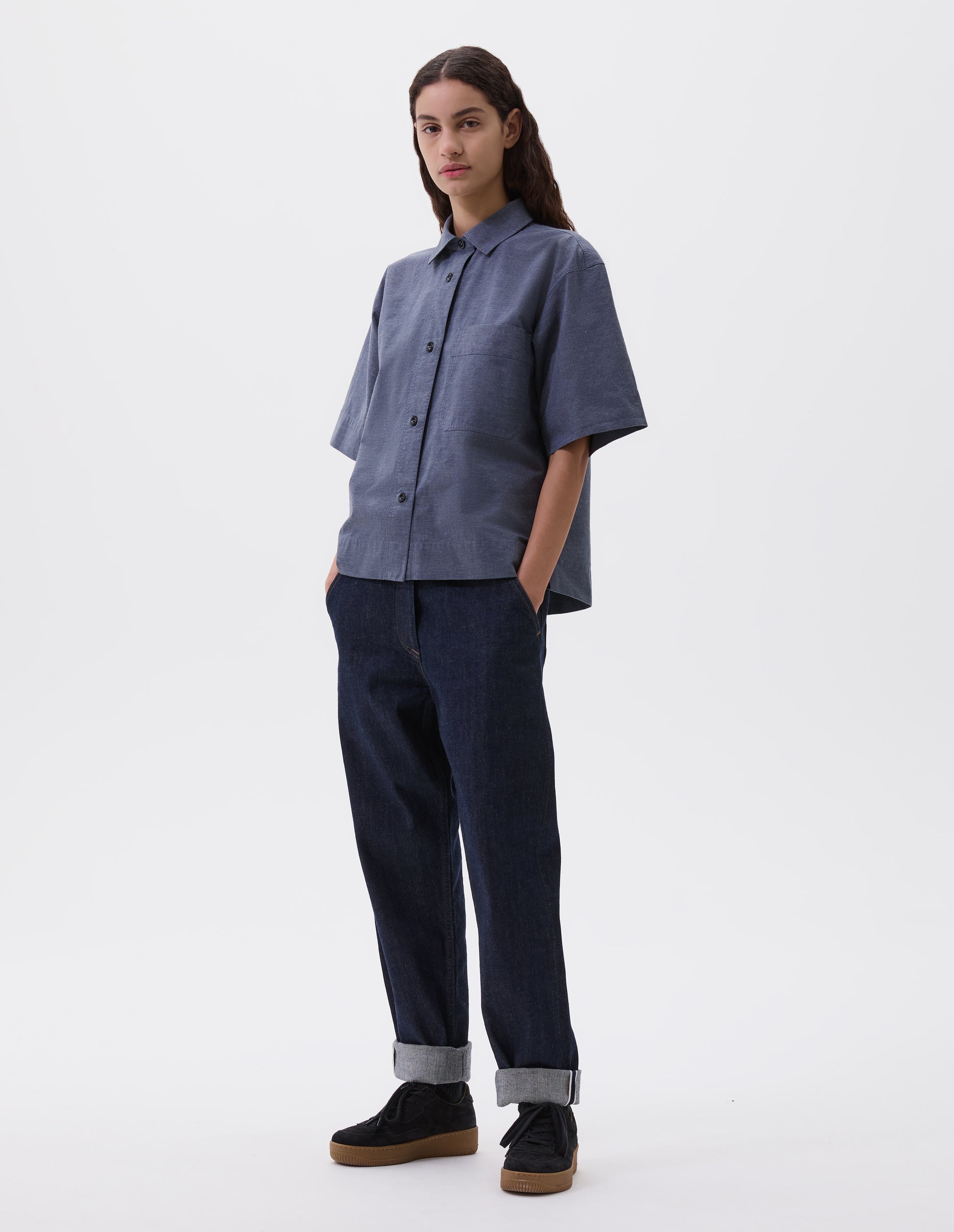Full body image, front view of model wearing  relaxed dark blue jeans with slant side pockets. Styled with blue short sleeve shirt and black nubuck trainers.