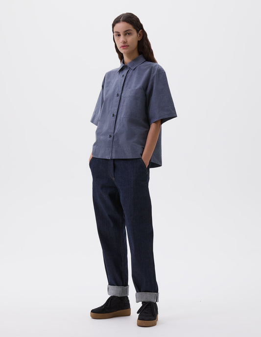 Full body image, front view of model wearing  relaxed dark blue jeans with slant side pockets. Styled with blue short sleeve shirt and black nubuck trainers.