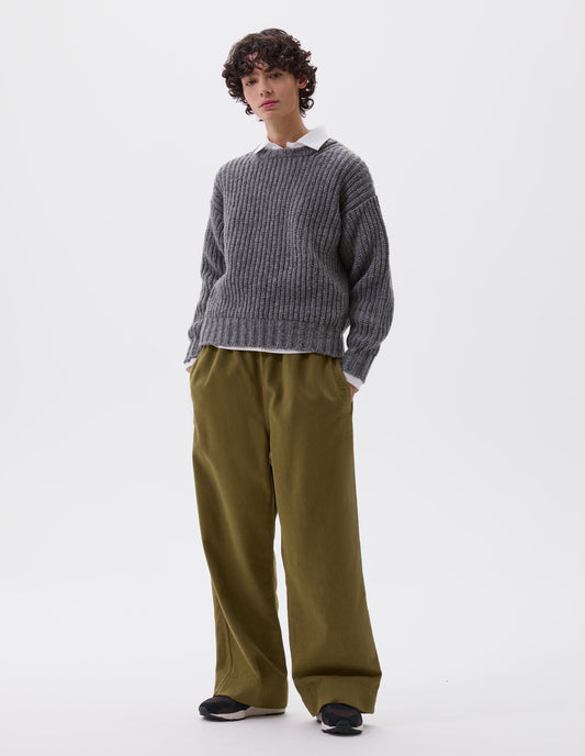 Full body image, front view of model wearing grey crew neck jumper with contrasting rib on neck, cuffs and hem. Styled over a white shirt, with green trousers and black and brown trainers.