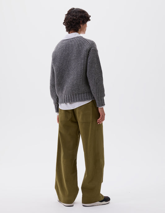 Full body image, back view of model wearing grey crew neck jumper with contrasting rib on neck, cuffs and hem. Styled over a white shirt, with green trousers and black and brown trainers.