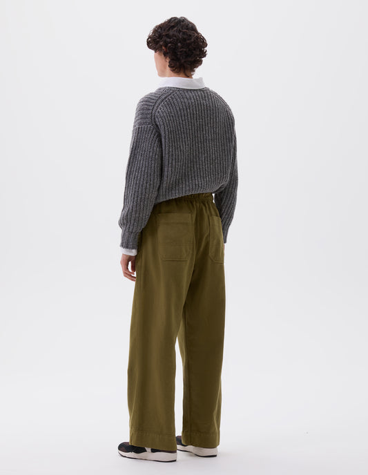 Full body image, back view of model wearing green relaxed wide leg joggers with elasticated waistband and patch back pockets. Styled with grey jumper over white shirt and black, brown and white trainers.