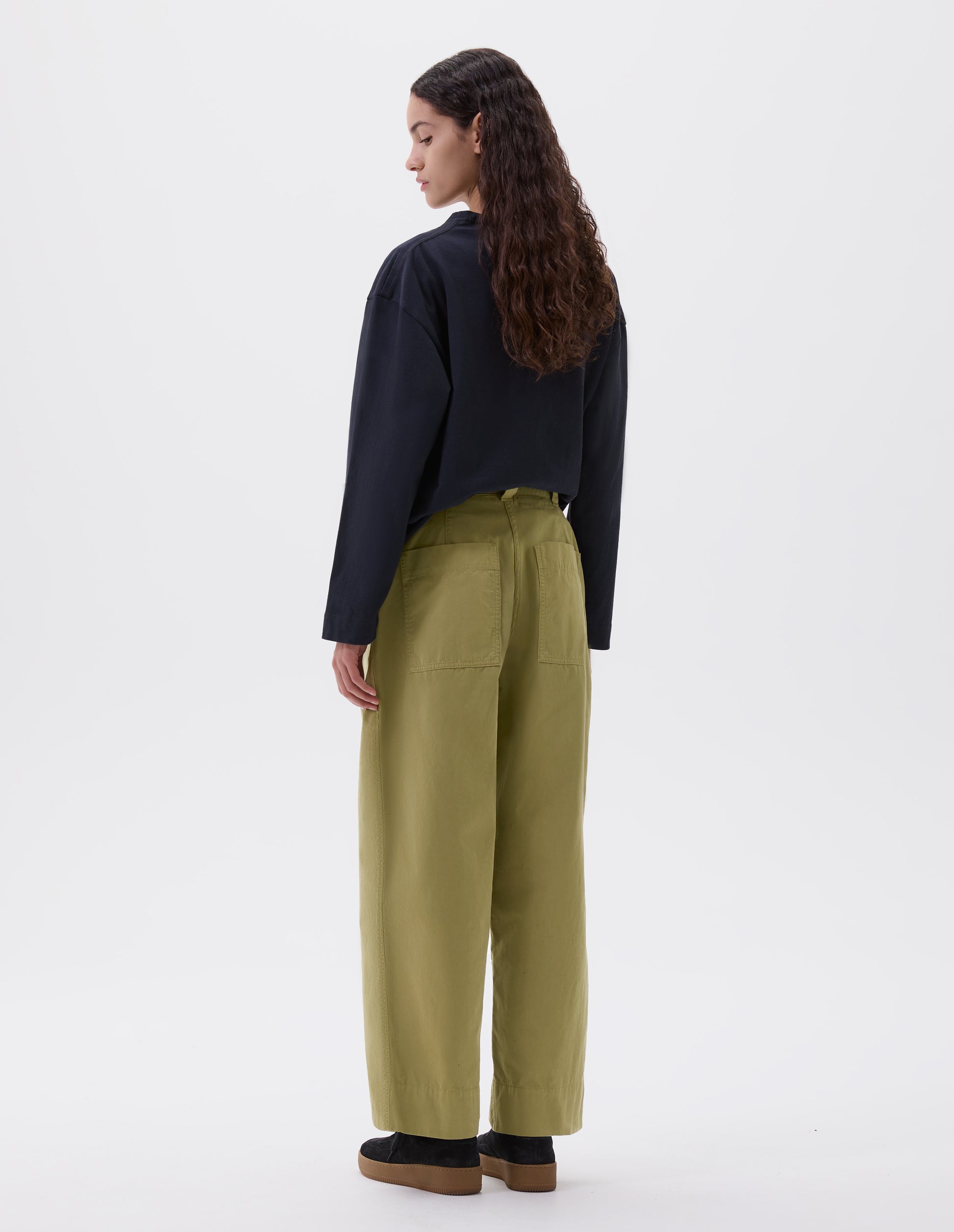 Full body image, back view of model wearing green trousers with  patch back pockets and deep stitch hem. Styled with navy blue long sleeve top and black suede trainers.