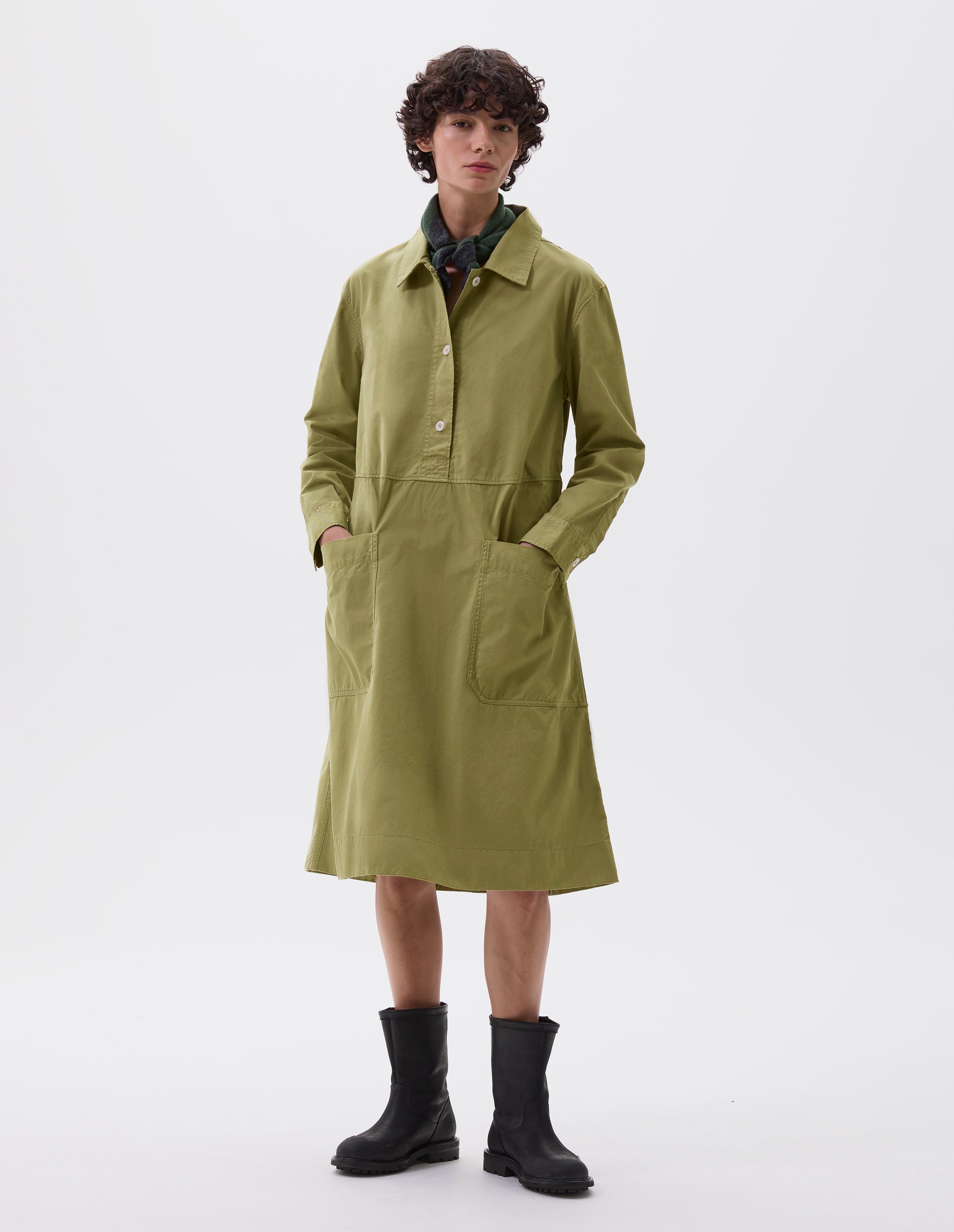 Full body image, front view of model wearing green long sleeve shirt dress with box stitch detailing, large apron patch pockets, deep stitch hem and waist seam detail. Styled with black leather biker boots and green knitted scout scarf.