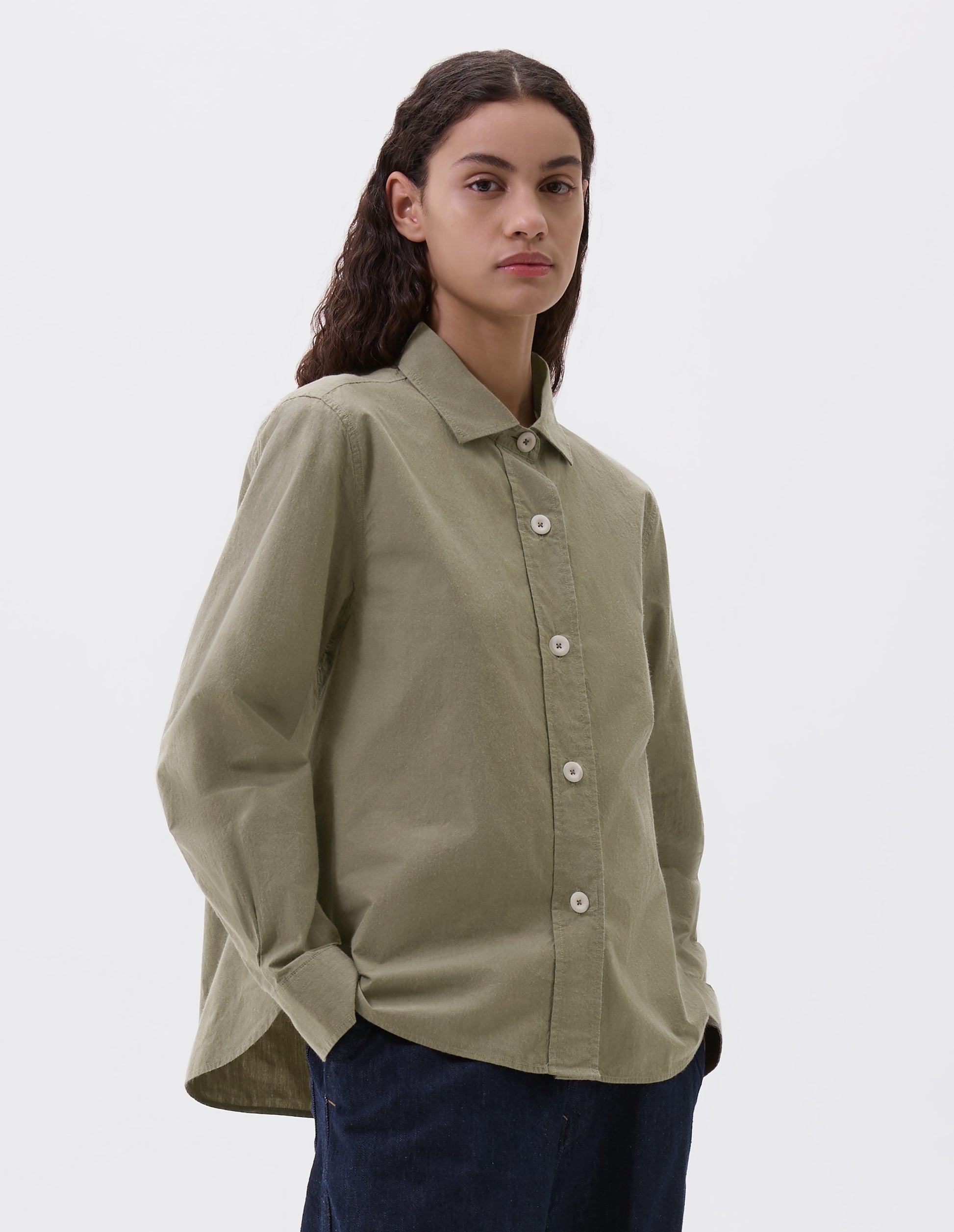 Cropped body image, front view of model wearing green brown long sleeve shirt, with stitch through raised placket, corozo button closure and cuffs and curved hem. Styled with indigo blue jeans.