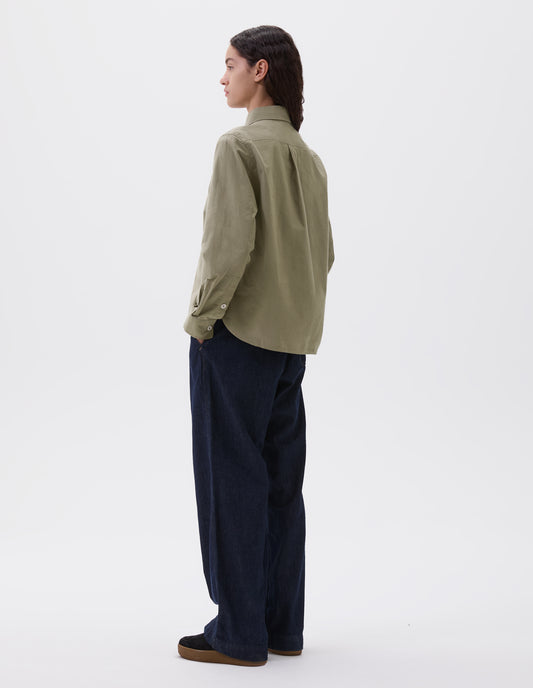 Full body image, back view of model wearing green brown long sleeve shirt, with box pleat on back yoke, corozo button cuffs and curved hem. Styled with indigo blue denim jeans and black suede trainers.