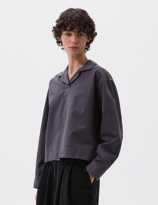 Cropped image, front view of model wearing greyish blue cropped open neck pull-on shirt with box stitch detailing on placket. Styled with black skirt.