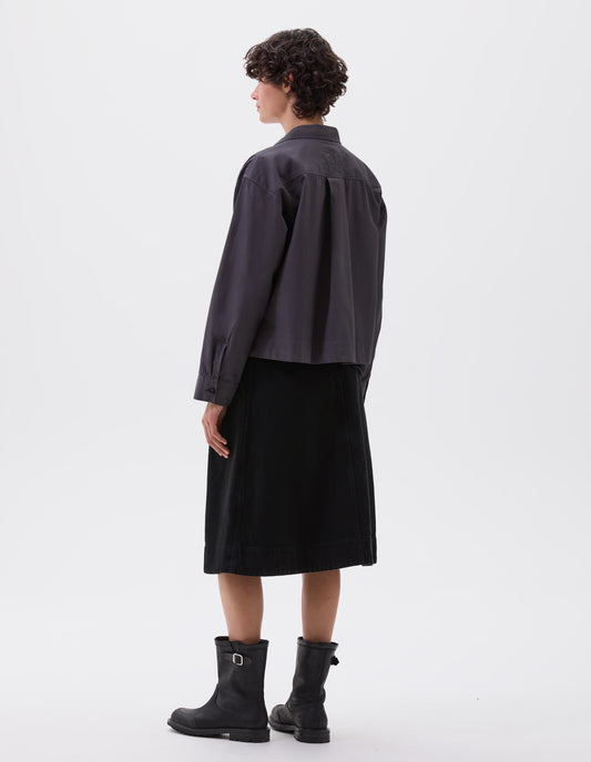 Full body image, back view of model wearing greyish blue cropped shirt with wide box pleat on back yoke and corozo brace buttons on cuffs. Styled with black skirt and black leather biker boots.