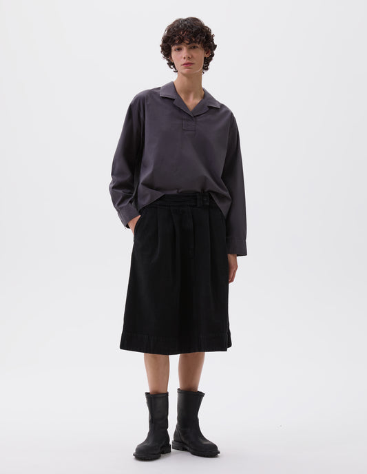 Full body image, front view of model wearing greyish blue cropped open neck pull-on shirt with box stitch detailing on placket. Styled with black skirt and black leather biker boots.