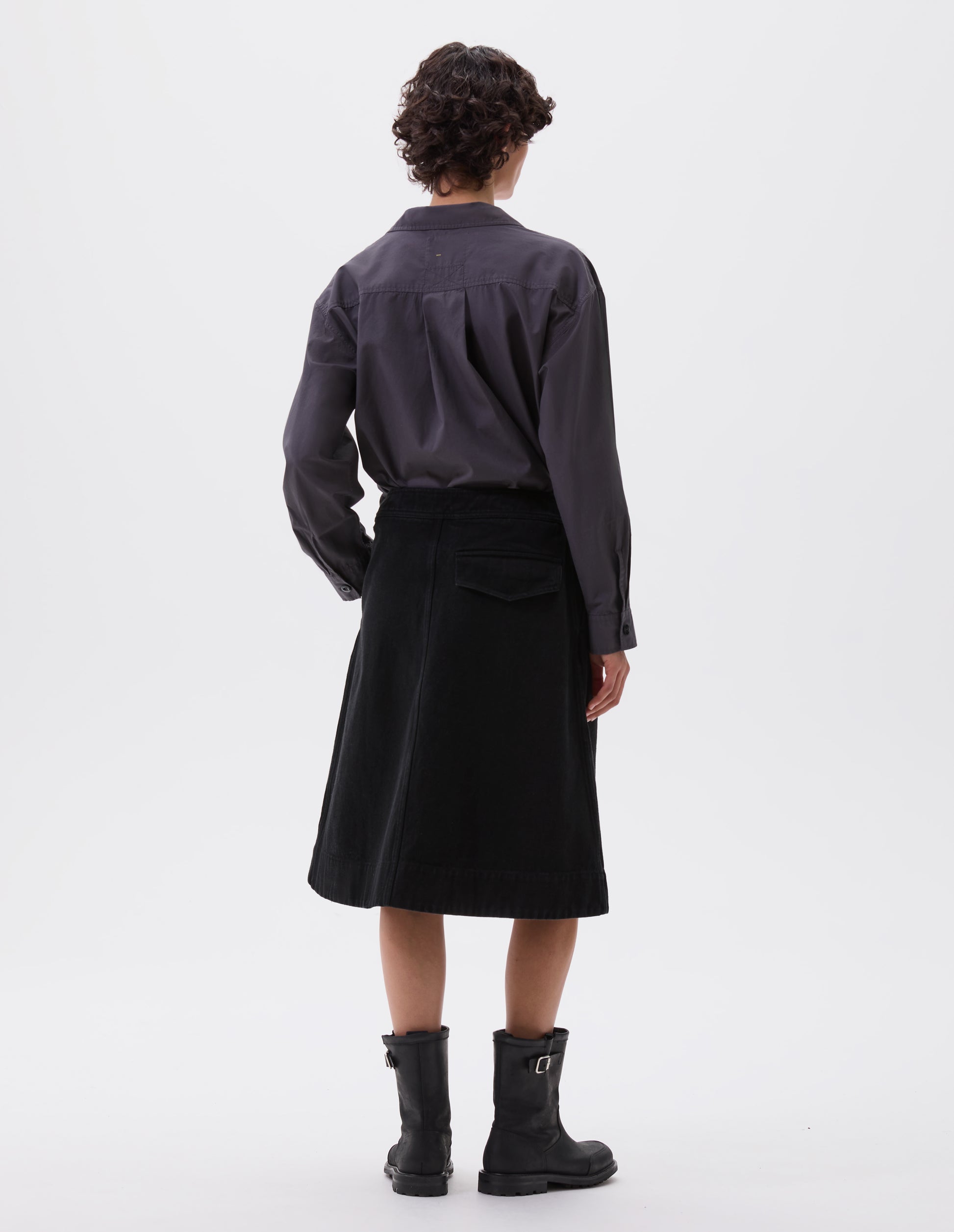 Full body image, back view of model wearing black A-line skirt with deep stitch hem and back flap pocket. Styled with grey pullover shirt and black leather biker boots.
