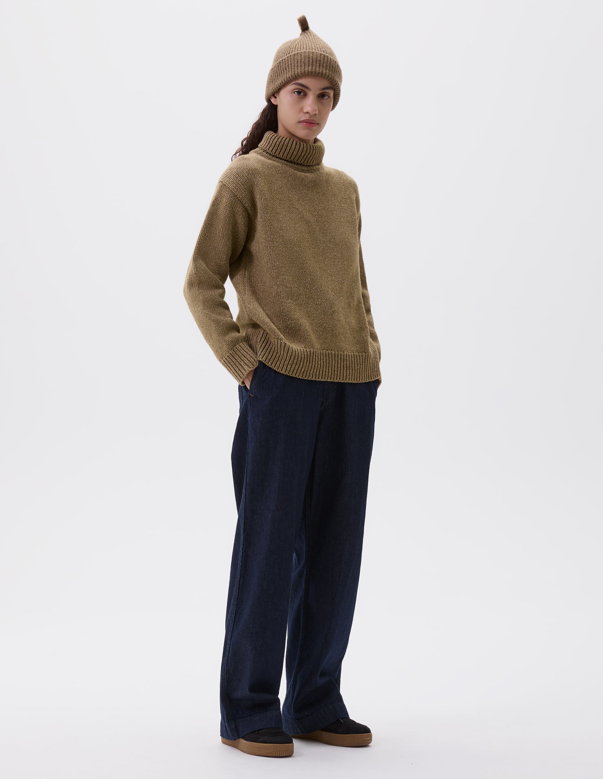 Full body image, front view of model wearing relaxed light green high neck jumper with deep rib trim on neck, cuffs and hem. Styled with light green beanie, dark blue jeans and black nubuck trainers. 