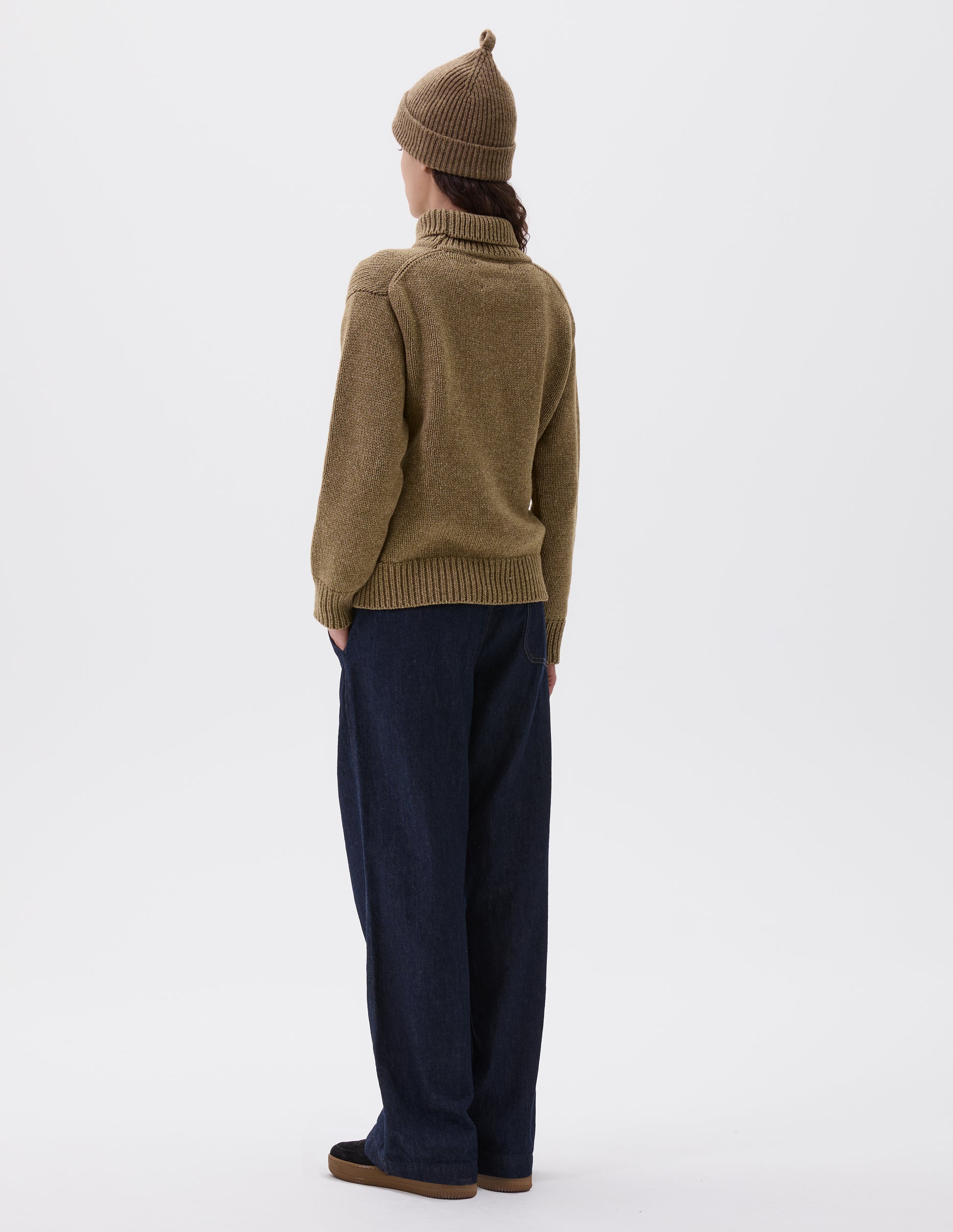 Full body image, back view of model wearing relaxed light green high neck jumper with deep rib trim on neck, cuffs and hem. Styled with light green beanie, dark blue jeans and black nubuck trainers. 
