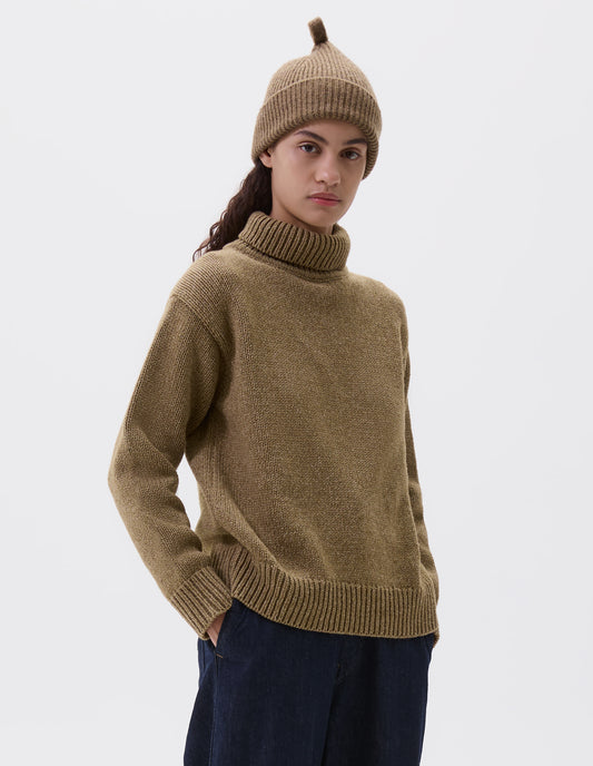 Cropped body image, front view of female model wearing greenish yellow wool ribbed hat, with knitted loop detailing on top. Styled with matching jumper and indigo blue jeans.