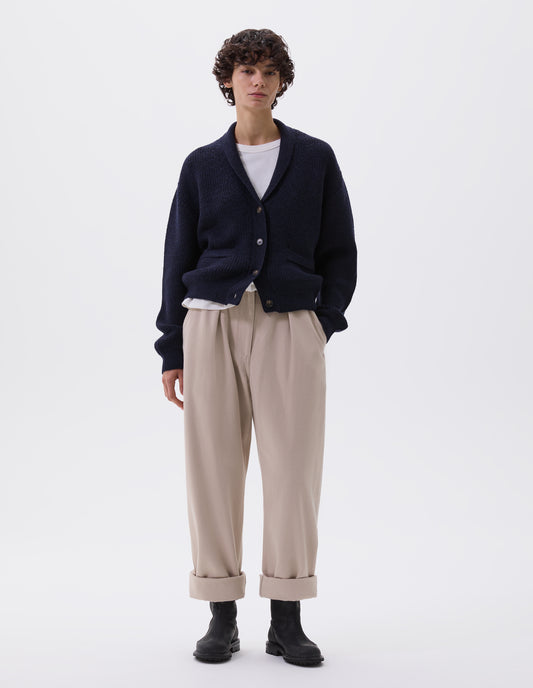 Full body image, front view of model wearing dark blue cardigan with ribbed collar, cuffs and hem, patch pockets and corozo brace buttons. Styled over white t-shirt, beige trousers and black leather biker boots.