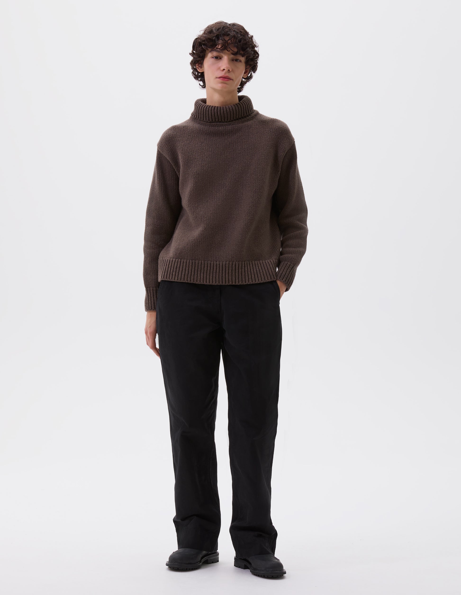 Full body image, front view of model wearing relaxed brown high neck jumper with deep rib trim on neck, cuffs and hem. Styled with black trousers and black leather boots.