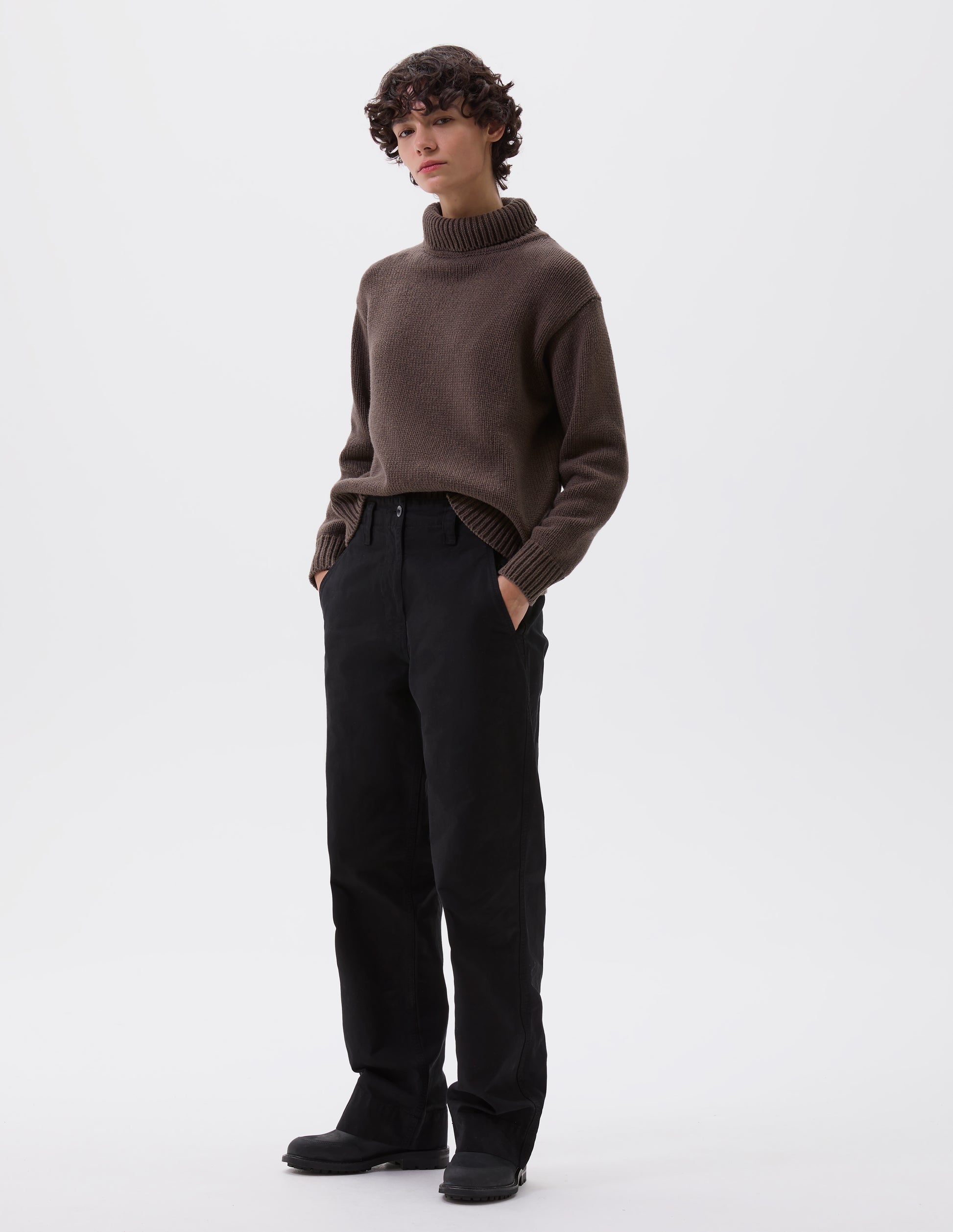 Full body image, front view of model wearing black cotton canvas trousers with stitch through waistband, button fastening, dropped belt loops and concealed slant pockets. Styled with brown high neck jumper and black leather boots.