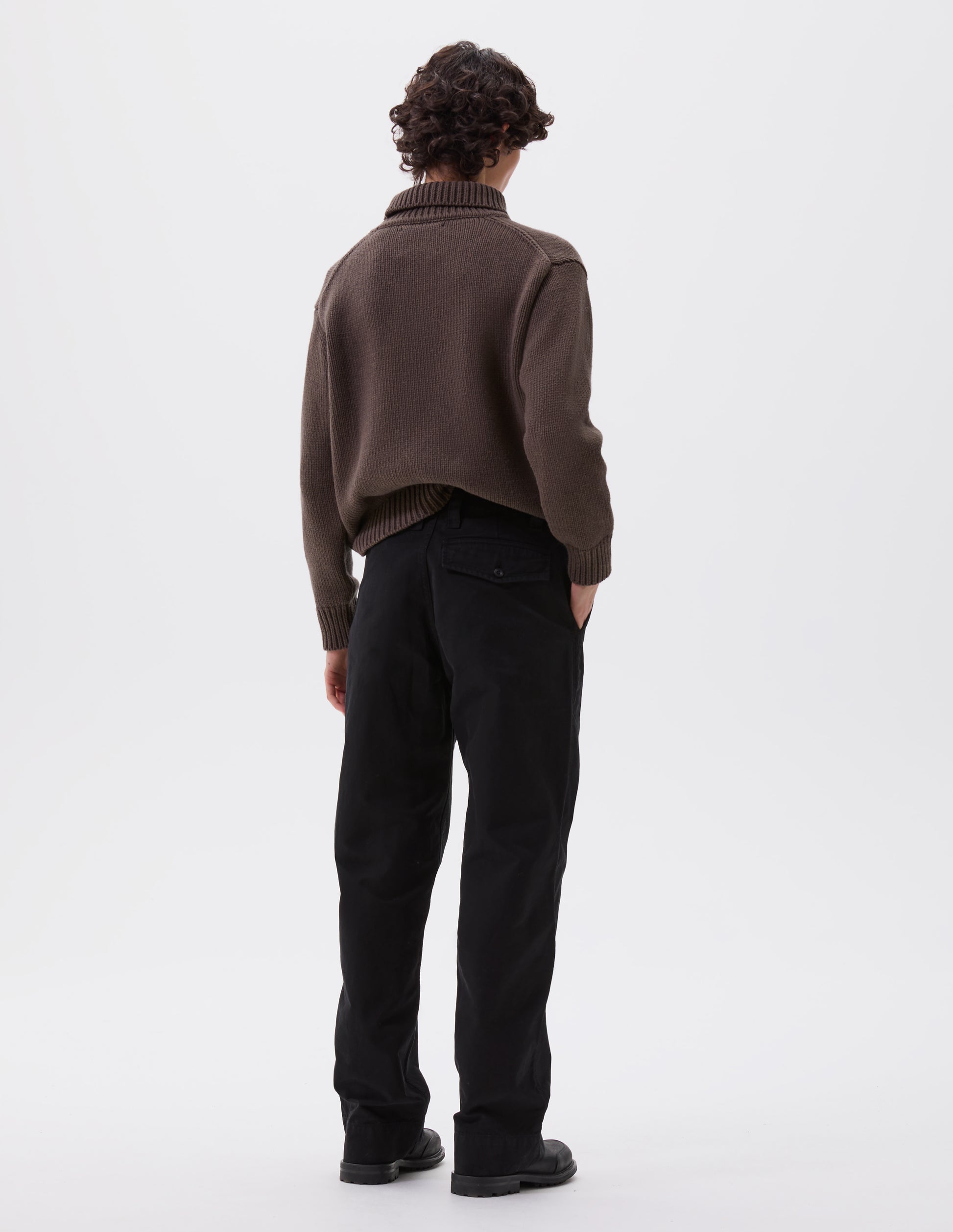 Full body image, back view of model wearing black cotton canvas trousers with stitch-through waistband, dropped belt loops and button through flap back pocket. Styled with brown high neck jumper and black leather boots.
