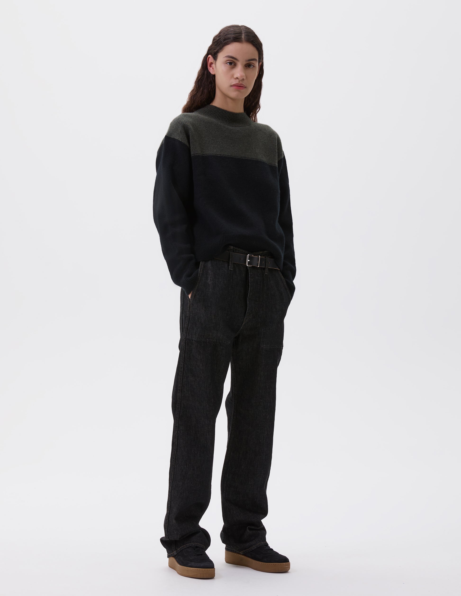 Full body image, front view of model wearing black denim twill jeans, with metal button fly, stitch-through waistband with belt loops, and stitch through pockets. Styled with green and black jumper, black leather belt and black suede trainers. 