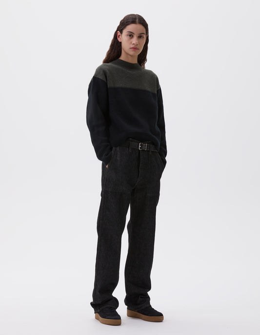 Full body image, front view of model wearing black denim twill jeans, with metal button fly, stitch-through waistband with belt loops, and stitch through pockets. Styled with green and black jumper, black leather belt and black suede trainers. 