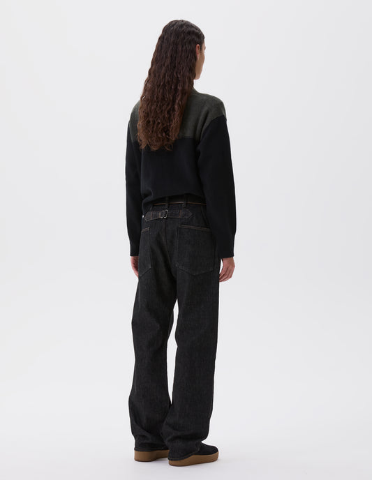 Full body image, back view of model wearing black denim twill jeans, with stitch-through waistband with belt loops, cinch back detail with metal buckle and patch back pockets. Styled with green and black jumper, black leather belt and black suede trainers. 
