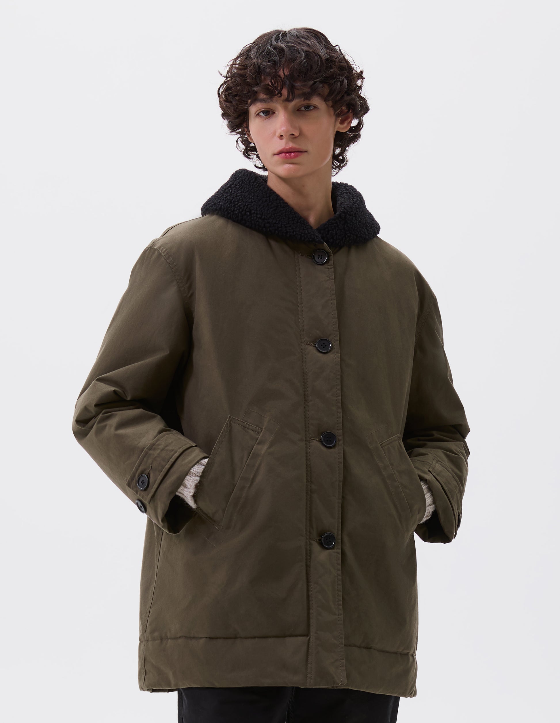 Cropped image, front view of model wearing relaxed khaki green coat with black fleece collar, slanted welt pockets, corozo brace buttons and button tab cuff adjusters. Styled with black trousers.
