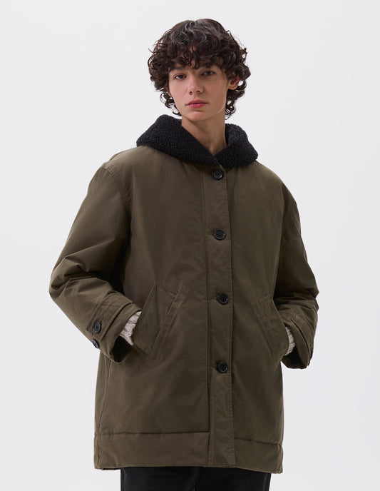 Cropped image, front view of model wearing relaxed khaki green coat with black fleece collar, slanted welt pockets, corozo brace buttons and button tab cuff adjusters. Styled with black trousers.