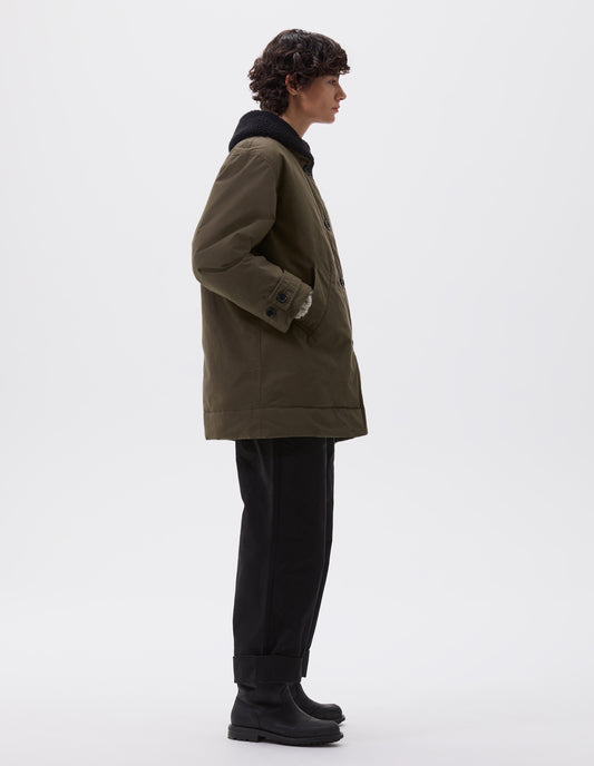 Full body image, side view of model wearing relaxed khaki green coat with black fleece collar, slanted welt pockets, corozo brace buttons and button tab cuff adjusters. Styled with black trousers and black leather boots.