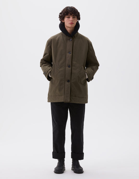 Full body image, front view of model wearing relaxed khaki green coat with raised black fleece collar, slanted welt pockets, corozo brace buttons and button tab cuff adjusters. Styled with black trousers and black leather boots.