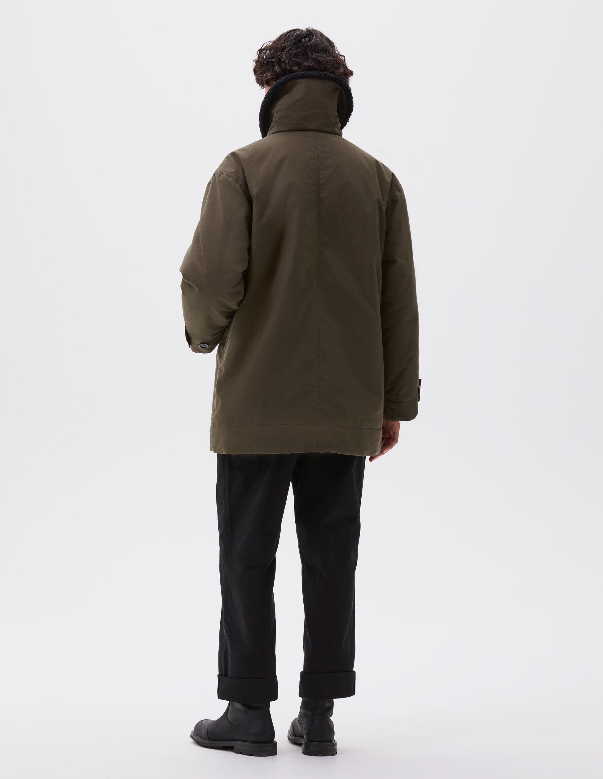 Full body image, back view of model wearing relaxed khaki green coat with raised black fleece collar and corozo brace button tab cuff adjusters. Styled with black trousers and black leather boots.