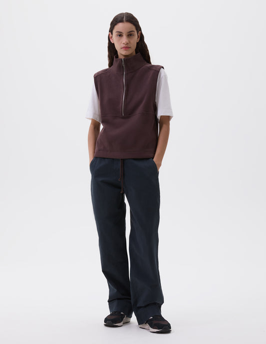 Full body image, front view of model wearing purple brown half zip high neck sleeveless track top and drawcord hem. Styled over white t-shirt, dark blue trousers with side pockets and black, brown and white trainers. 