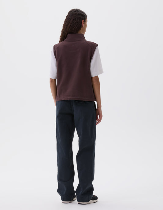 Full body image, back view of model wearing purple brown high neck sleeveless track top. Styled over white t-shirt, dark blue trousers and black, brown and white trainers. 