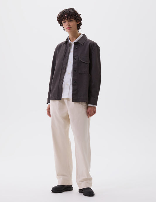 Full body image, front view of model wearing grey oversized shirt with stitch through raised placket, corozo button fastening, patch chest pocket and deep stitch hem. Styled over white cotton shirt with off white trousers and black leather boots.