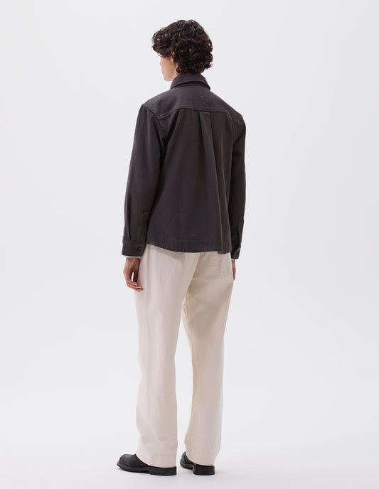 Full body image, back view of model wearing grey oversized shirt with corozo button fastening cuffs, box pleat on back yoke and deep stitch hem. Styled with off white trousers and black leather boots.