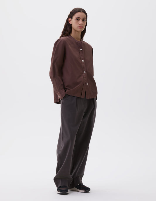 Full body image, front view of model wearing brown purple collarless long sleeve shirt with raised placket and corozo button closure. Styled with brown pleated trousers and brown and black trainers. 