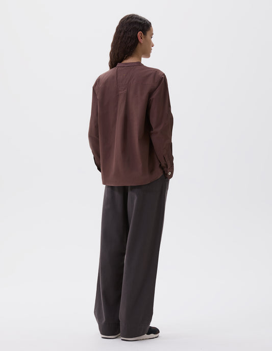 Full body image, back view of model wearing brown purple collarless long sleeve shirt with corozo button cuffs. Styled with  brown trousers and brown and black trainers. 