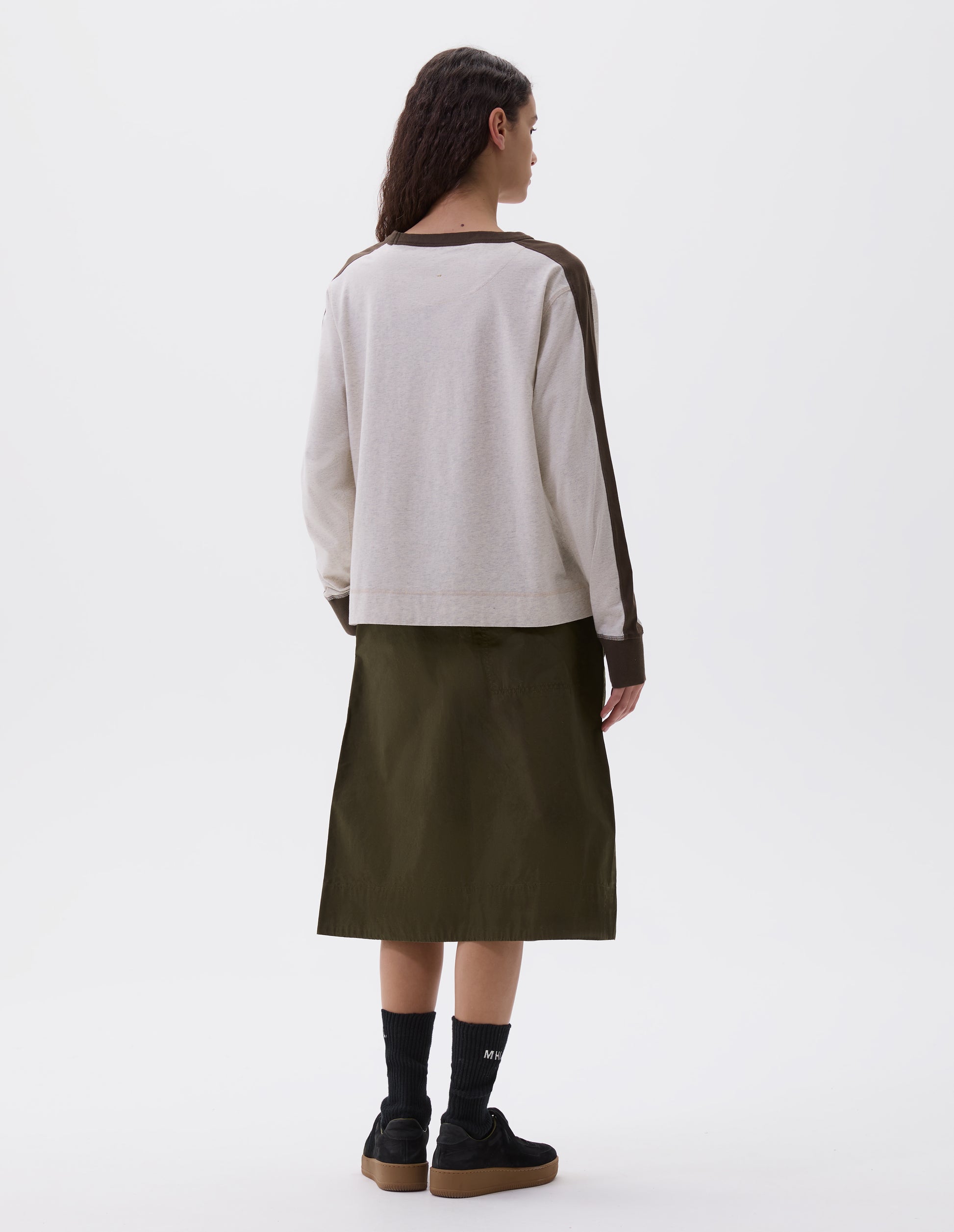 Full body image, back view of model wearing white long sleeve t-shirt with contrasting green stripe on cuffs, arms and neckline. Styled with green skirt and black socks and black nubuck trainers.