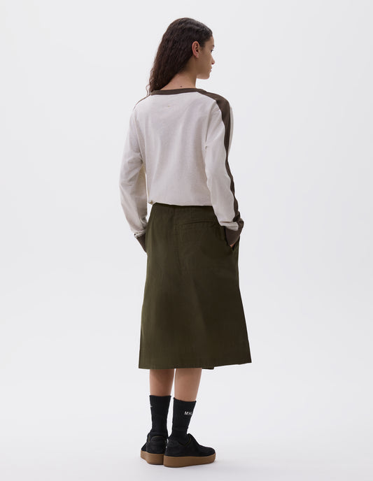 Full body image, back view of model wearing green skirt with deep wrap and slant side pockets. Styled with beige and green long sleeve sweatshirt, black socks and black nubuck leather trainers.