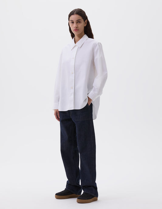 Full body image, front view of model wearing white long sleeve cotton shirt, with corozo button closure and cuffs and elongated hem. Styled with indigo blue jeans and black nubuck trainers.
