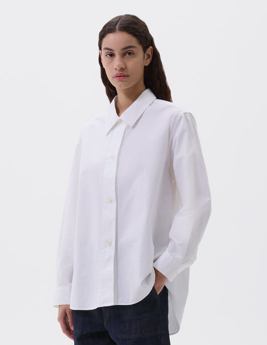 Cropped body image, front view of model wearing white long sleeve cotton shirt, with corozo button closure and cuffs and elongated hem. Styled with indigo blue jeans.