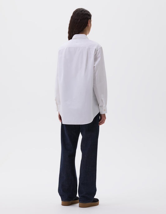 Full body image, back view of model wearing white long sleeve cotton shirt, with corozo cuffs, elongated hem and pleated back yoke. Styled with indigo blue jeans and black nubuck leather trainers.