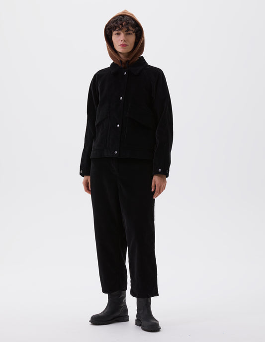 Full body image, front view of model wearing black heavy corduroy raglan sleeve jacket, with press stud fastening offset placket, slant patch pockets with storm flap closure and adjustable cuffs. Styled with brown knitted hood, matching black corduroy trousers and black leather boots.