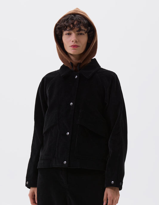 Cropped body image, front view of model wearing black heavy corduroy raglan sleeve jacket, with press stud fastening offset placket, slant patch pockets with storm flap closure and adjustable cuffs. Styled with brown knitted hood and matching black corduroy trousers.