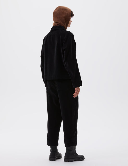 Full body image, back view of model wearing black heavy corduroy raglan sleeve jacket, with press stud fastening adjustable cuffs. Styled with brown knitted hood, matching black corduroy trousers and black leather boots.