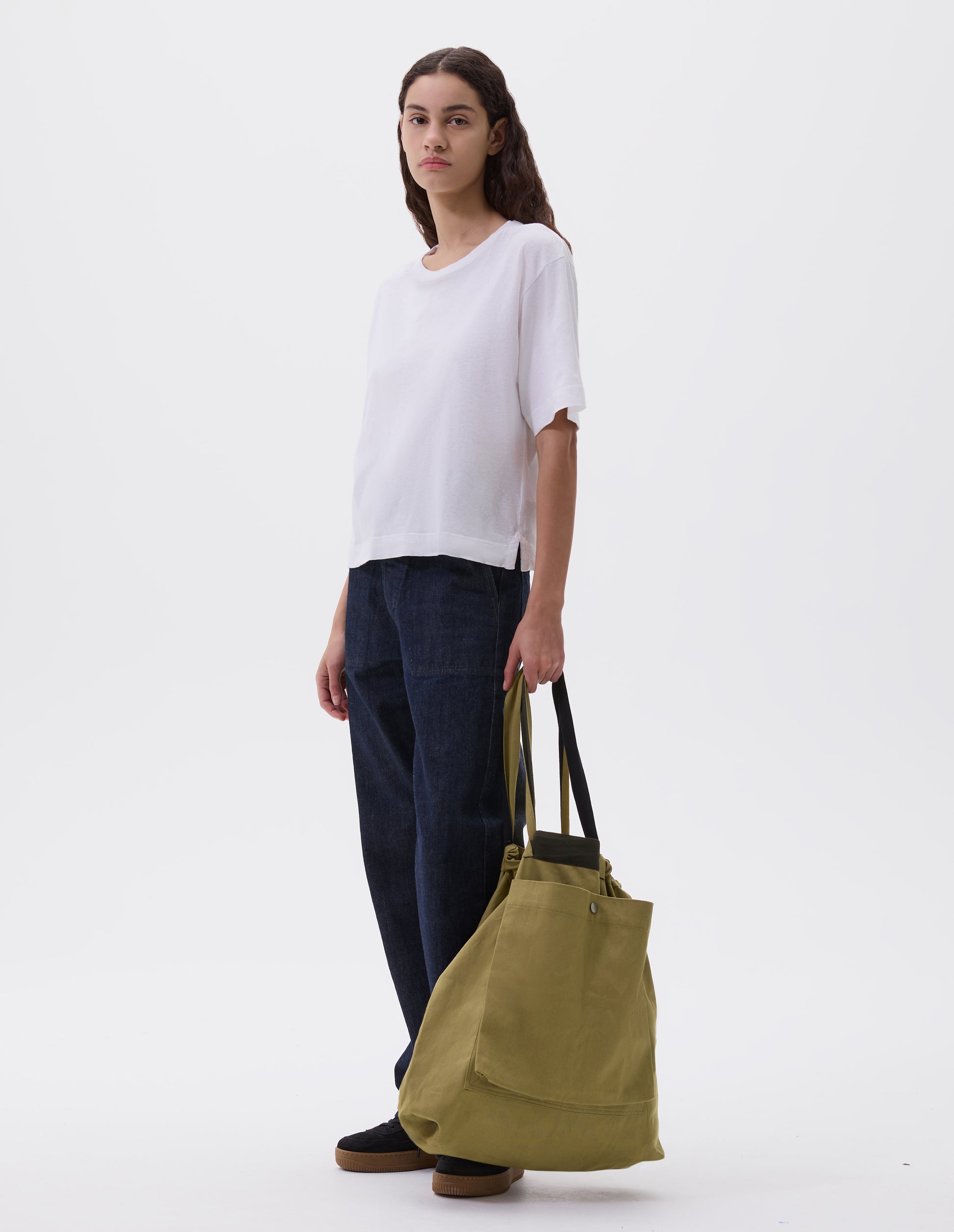 Full body image, front view of model holding olive green tote bag with drawcord handles, large press stud fastening bellows patch pocket and contrasting herringbone tape detailing. Styled with white t-shirt, indigo blue denim jeans and black leather trainers.