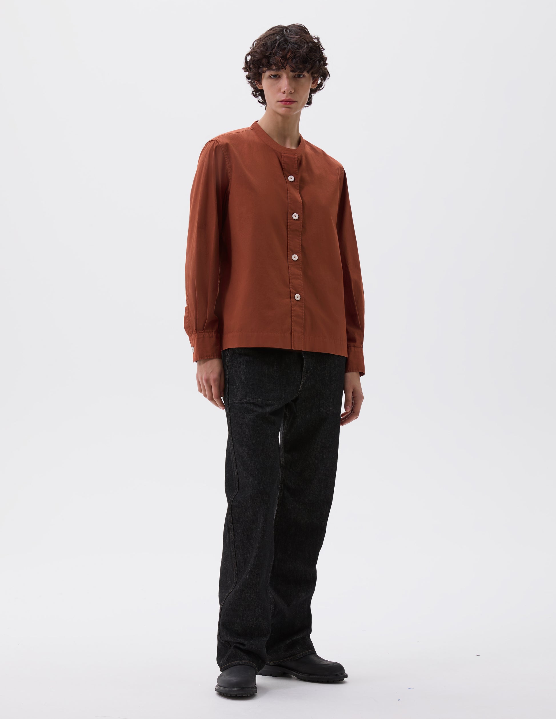 Full body image, front view of model wearing  brownish red collarless shirt with raised stitch through placket and corozo brace buttons. Styled with black denim jeans and black leather boots.  