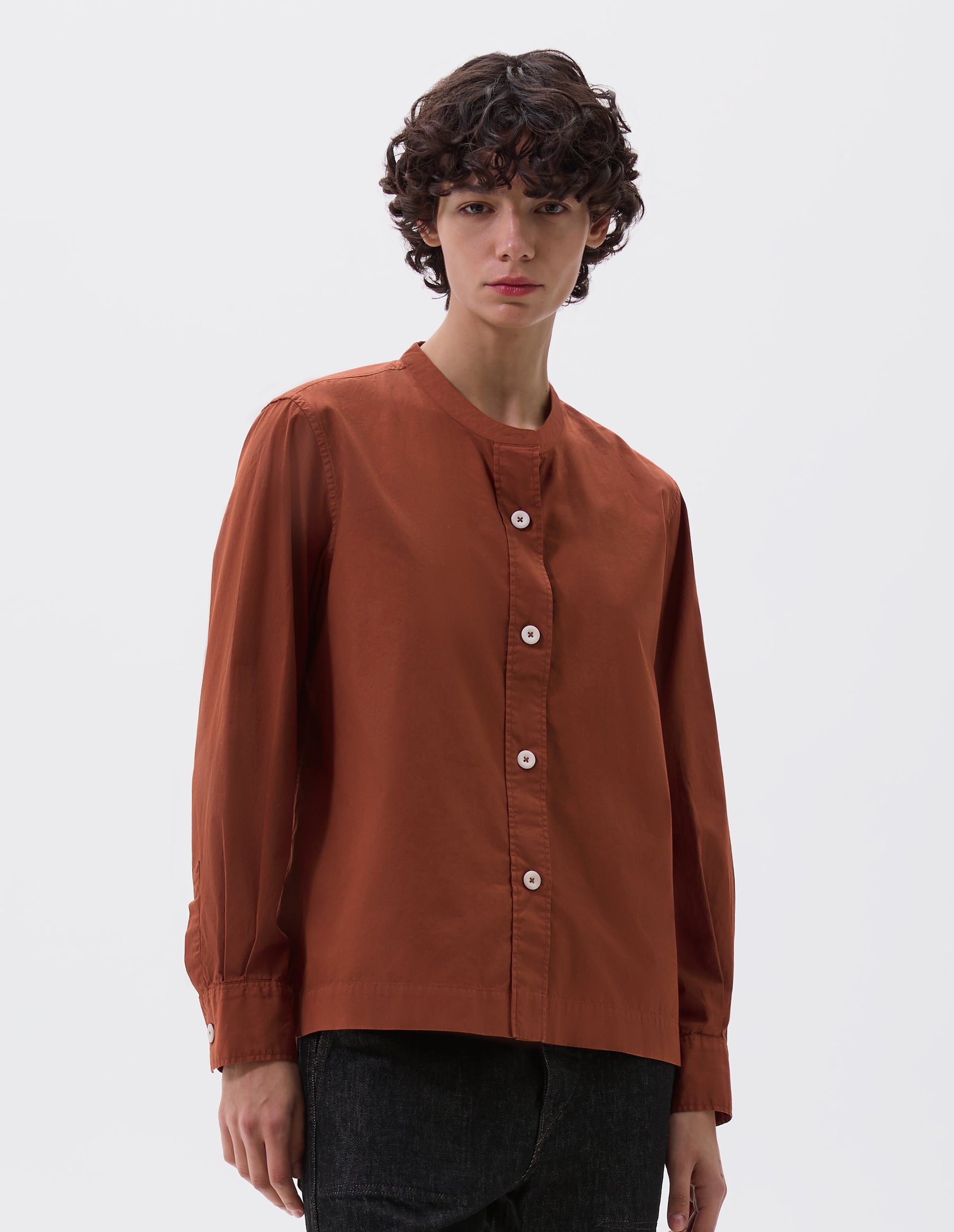 Cropped body image, front view of model wearing brownish red collarless shirt with raised stitch through placket and corozo brace buttons. Styled with black denim jeans. 