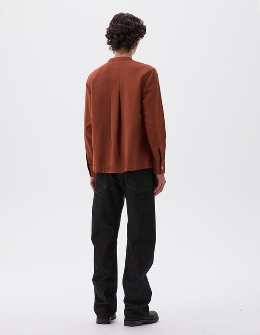 Full body image, back view of model wearing brownish red collarless shirt with corozo brace button cuffs and buttonhole detailing on neck band and box pleat on back yoke. Styled with black denim jeans and black leather boots.  