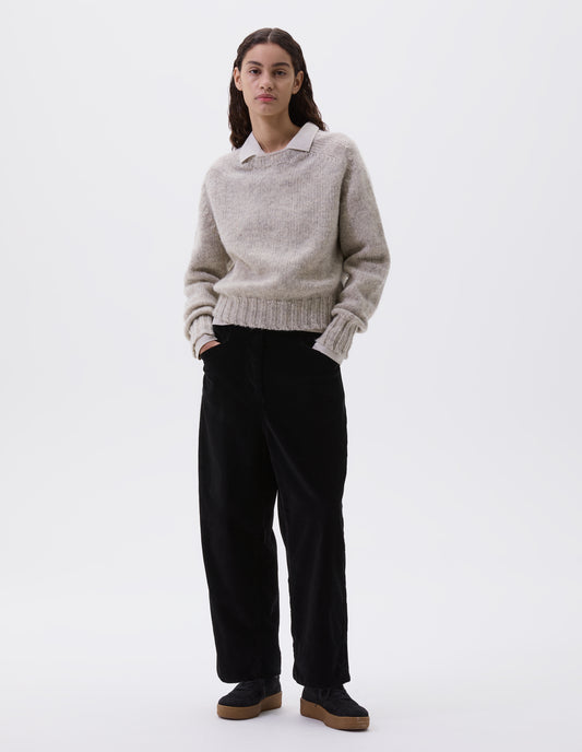 Full body image, front view of model wearing black heavy corduroy trousers with concealed double hook and bar fastening, frog pockets and deep stitch-through hem. Styled with off white shirt, beige jumper and black suede trainers.