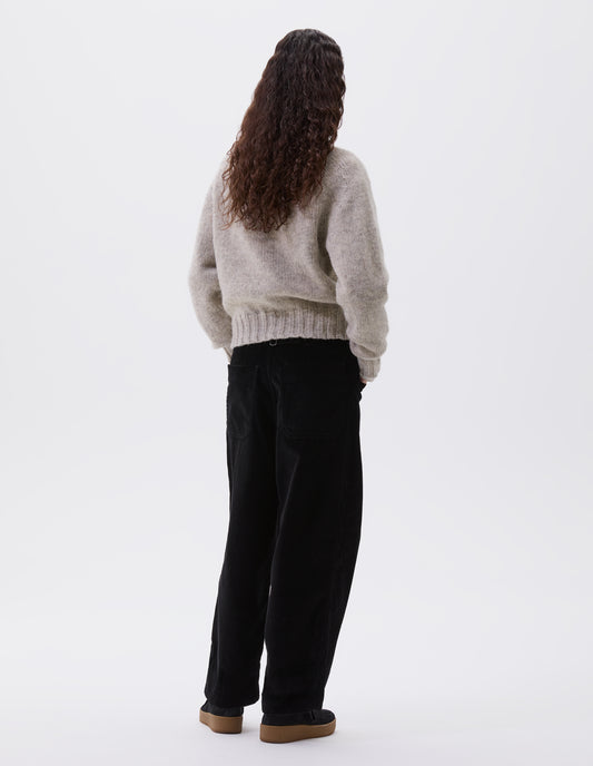 Full body image, back view of model wearing black heavy corduroy trousers with frog pockets, deep stitch-through hem, patch back pockets and cinch waist adjusters. Styled with beige jumper and black suede trainers.