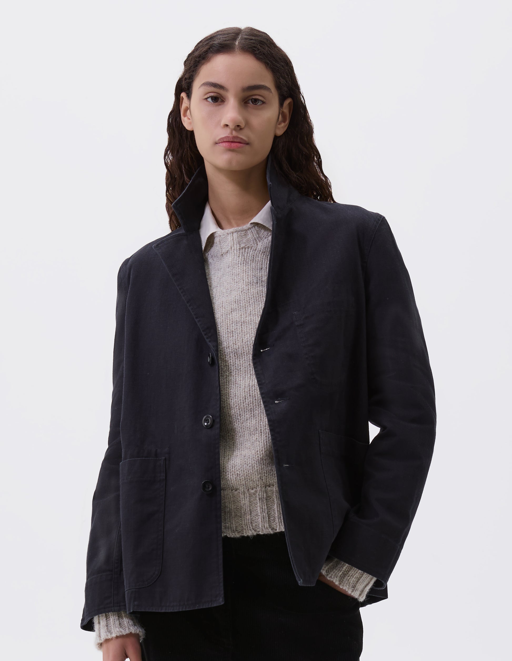 Cropped body image, front view of model wearing indigo twill single breasted blazer, with two front patch pockets, fold back split cuff, curved hem and corozo button closure. Styled over beige shirt and beige jumper and black trousers.